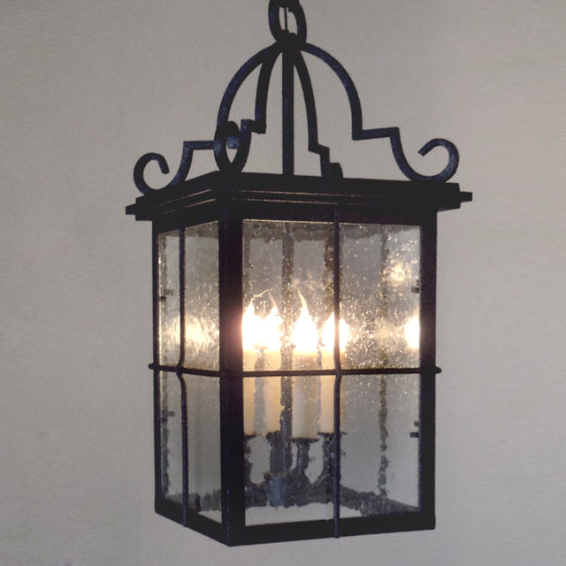 A intricately designed wrought iron pendant light featuring a classic lantern shape. Four candle-style bulbs illuminate through textured glass panels, framed by ornate detailing. The piece complements a variety of decor styles while providing warm, inviting light.