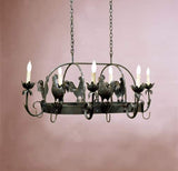 A large wrought iron chandelier featuring intricate rooster motifs, elegantly suspended from a chain. It has six candle-like lights, surrounded by decorative floral elements. The design reflects a rustic charm, perfect for adding character to any space.