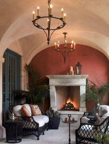 Two elegant wrought iron chandeliers hang from a curved ceiling, illuminating a cozy seating area beside a stone fireplace. The warm ambiance is enhanced by plush seating and decorative elements, creating a welcoming and sophisticated space.