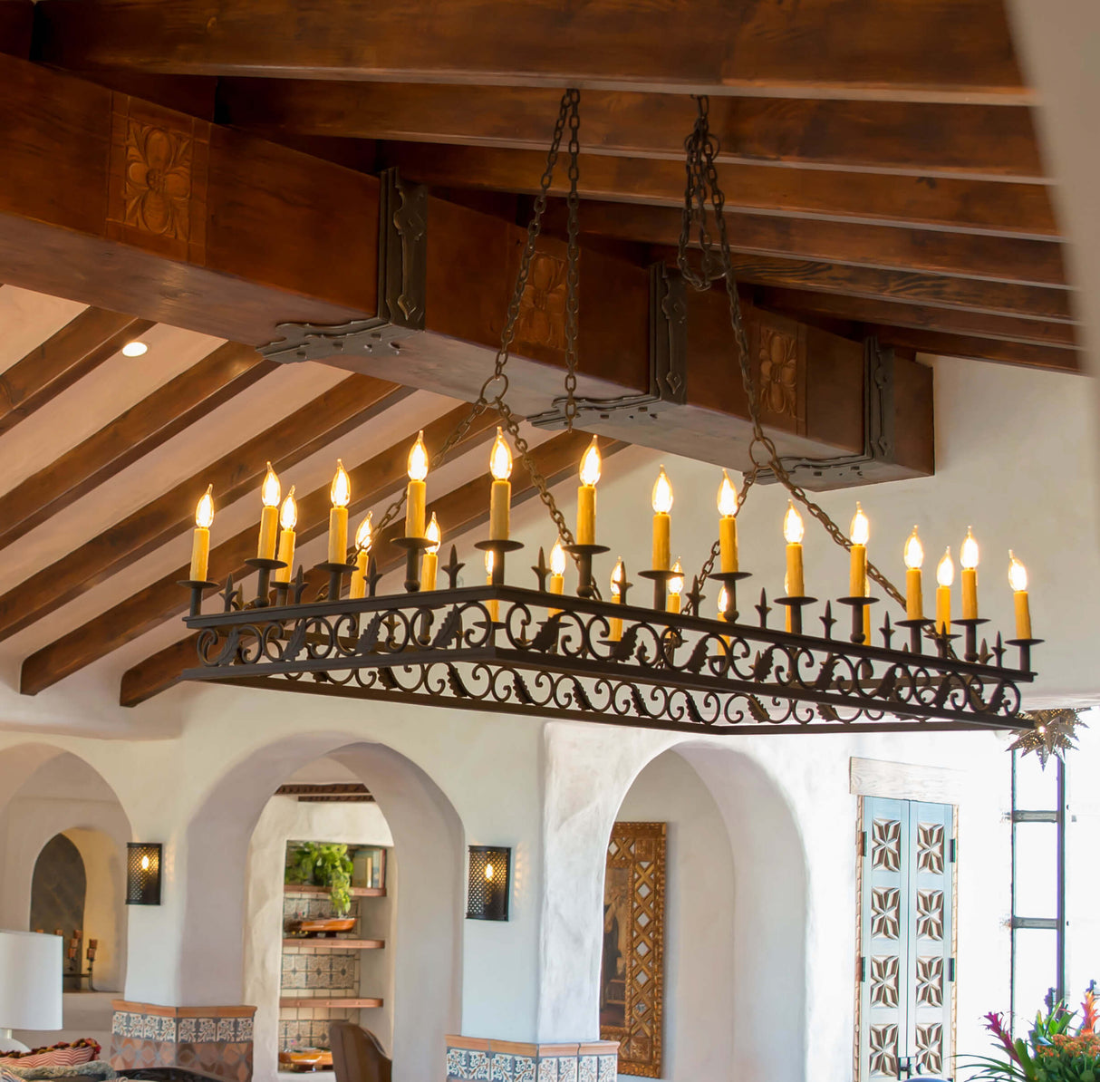 A large, ornate chandelier featuring multiple candle-like bulbs rests from a chain, enhancing the rustic, warm ambiance of a ceiling with wooden beams. Its intricate wrought iron design adds character to the space, surrounded by white plaster walls and decorative accents.