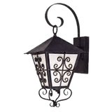 A decorative wall sconce featuring a hexagonal design with a pointed roof. The fixture showcases intricate scrollwork in black wrought iron and frosted glass panels, creating a warm, inviting glow for exterior or interior spaces.