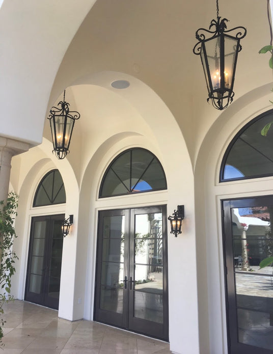 Two elegant wrought iron sconces illuminate a grand entryway with arched doorways and large glass doors. The warm glow enhances the inviting atmosphere, showcasing the intricate designs of the fixtures against a light-colored wall. The sconces complement the architectural features, while lush greenery adds a touch of nature to the scene.