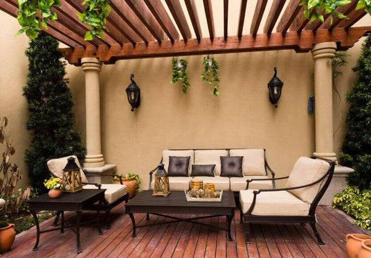 A beautifully designed outdoor seating area features a wrought iron wall sconce, surrounded by lush greenery and warm wooden beams. The elegant furniture invites relaxation, while decorative lanterns add charm to the cozy ambiance.