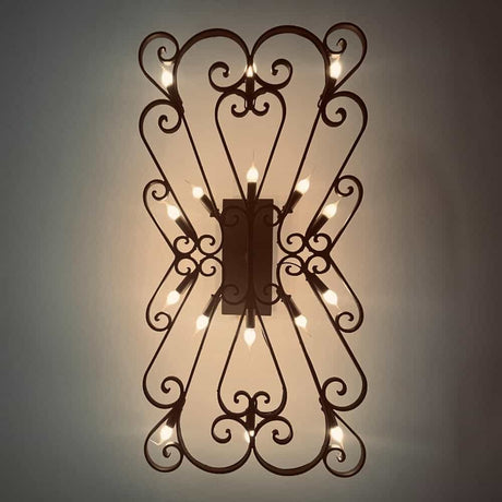 A decorative wall sconce featuring intricate wrought iron scrollwork and multiple candle-like light bulbs, casting a warm glow on a soft background.