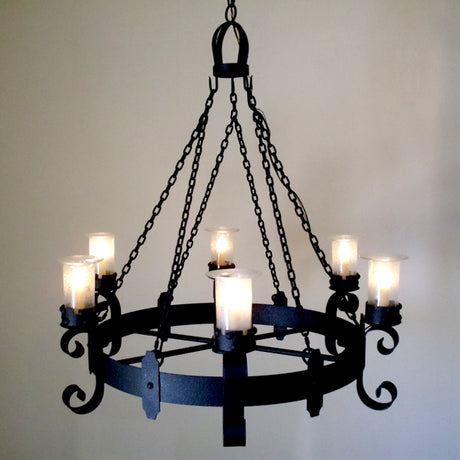 A wrought iron chandelier featuring a circular design with intricately forged arms. It showcases six glass candle holders with flickering candle-like lights, suspended from a decorative chain. The piece combines elegance and classic craftsmanship, adding warmth and character to any space.