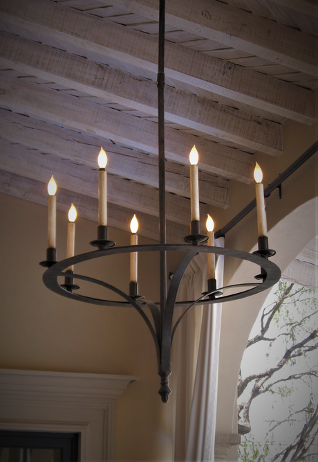 A striking wrought iron chandelier featuring a circular design with six candle-like lights. The fixture showcases a rustic aesthetic, complementing the wooden beams above. Ideal for adding warmth and character to any space.