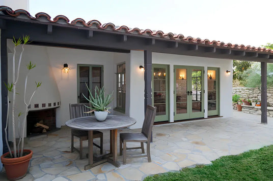 A charming outdoor patio featuring a rustic stone floor, a wooden table with chairs, and a potted plant. Elegant wrought iron sconces illuminate the space, enhancing the inviting atmosphere of this cozy seating area.