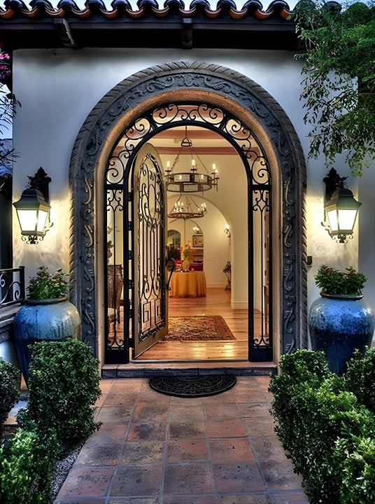 A beautifully designed entrance featuring a wrought iron door and decorative sconces. Inside, a chandelier hangs elegantly in a warm, inviting space, complemented by lush greenery and terracotta tiles. The entryway reflects craftsmanship and style, showcasing a blend of intricate ironwork and welcoming ambiance.